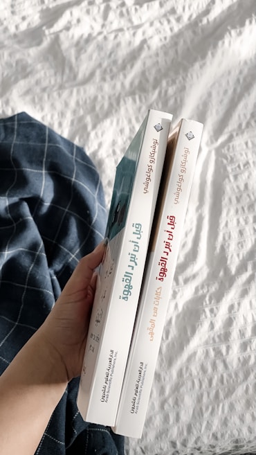 A hand holding two books with Arabic titles on the spines. The books are positioned vertically side by side, and are held over a white quilted fabric with a blue plaid cloth nearby.
