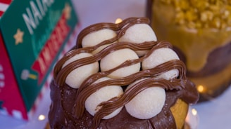 A dessert topped with six round white confections drizzled with chocolate, placed on a larger chocolate-covered pastry. In the background, a decorated card with festive colors and a secondary dessert topped with nuts are visible.