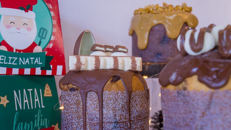 A festive arrangement featuring cakes with rich chocolate drizzle and decorative toppings. Two holiday-themed boxes with Christmas designs, including Santa Claus and inscriptions in Portuguese, accompany the desserts.