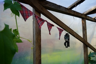 A rustic greenhouse interior with wooden beams and a makeshift flag banner made of plaid fabric. A black vintage-style hanging lantern is also visible. Green leaves and tendrils creep along the wooden structure, adding a natural touch to the setting.