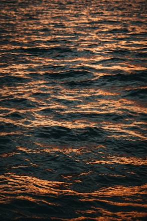 Dark ocean waves are illuminated by a warm orange glow, likely from a sunset or sunrise. The surface of the water has a textured and reflective appearance, with ripples and small waves creating a dynamic pattern across the scene.