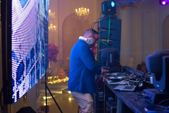 A DJ wearing headphones and a blue jacket is operating behind a mixing console at an event. The scene includes a large LED screen to the left and professional audio equipment around the DJ. The background features elegant chandeliers and subtle decorations, suggesting a formal gathering or party atmosphere.