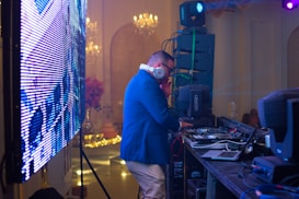 A DJ wearing headphones and a blue jacket is operating behind a mixing console at an event. The scene includes a large LED screen to the left and professional audio equipment around the DJ. The background features elegant chandeliers and subtle decorations, suggesting a formal gathering or party atmosphere.