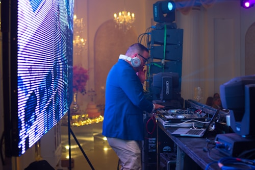 A DJ wearing headphones and a blue jacket is operating behind a mixing console at an event. The scene includes a large LED screen to the left and professional audio equipment around the DJ. The background features elegant chandeliers and subtle decorations, suggesting a formal gathering or party atmosphere.
