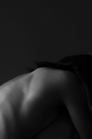 A black and white image captures the contours of a human back, slightly turned and bent forward. The skin is smooth and the play of light and shadow highlights the shoulder blades and spine, creating an artistic, intimate atmosphere.