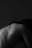 A black and white image captures the contours of a human back, slightly turned and bent forward. The skin is smooth and the play of light and shadow highlights the shoulder blades and spine, creating an artistic, intimate atmosphere.
