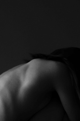 A black and white image captures the contours of a human back, slightly turned and bent forward. The skin is smooth and the play of light and shadow highlights the shoulder blades and spine, creating an artistic, intimate atmosphere.