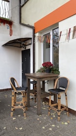 A small outdoor patio scene with a wooden table and two wicker bar stools. The area is decorated with potted red flowers and colorful hanging decorations. The walls are painted white with a distinctive orange strip near the top. Fallen leaves are scattered on the cobblestone ground.