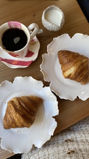 A cozy breakfast setup with two croissants on elegant white plates, a cup of black coffee in a colorful saucer, and a small speckled milk jug, all set on a wooden table.