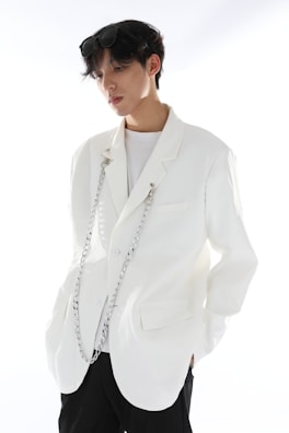 A person with short dark hair wearing a stylish white blazer adorned with decorative chains. The individual is also wearing sunglasses on their head and a plain white shirt underneath. The background is bright, enhancing the minimalist and modern fashion aesthetic conveyed.