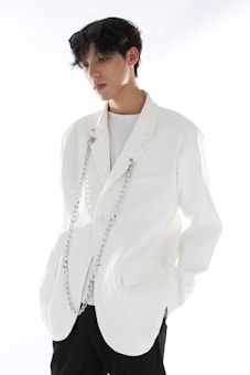 A person with short dark hair wearing a stylish white blazer adorned with decorative chains. The individual is also wearing sunglasses on their head and a plain white shirt underneath. The background is bright, enhancing the minimalist and modern fashion aesthetic conveyed.