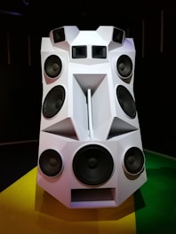 A large, futuristic speaker system with multiple speakers of various sizes arranged in a geometric, polygonal structure. The structure is predominantly white with angular shapes and black speaker cones. It is placed on a brightly colored floor, consisting of yellow and green sections.
