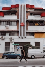 A street scene featuring a person walking past a white and brick building with red accents. The facade has multiple windows, balconies, and air conditioning units. Two vehicles, a blue car and a white van, are parked along the street. The building displays the number 82 on a red sign. A street sign reading 'Tiong Poh Rd' and a direction sign are also visible.