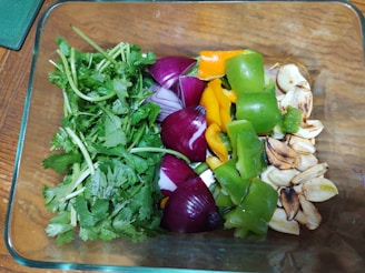 A glass container holds a variety of fresh vegetables, including coriander leaves, red onion pieces, green and yellow bell peppers, and roasted garlic cloves. The colorful assortment is neatly arranged on a wooden surface.