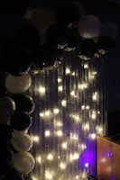 An arrangement of black and white balloons is displayed next to string lights hanging on a vertically striped backdrop. The lights emit a soft warm glow, highlighting the balloons and creating a festive atmosphere.