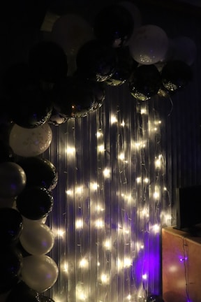 An arrangement of black and white balloons is displayed next to string lights hanging on a vertically striped backdrop. The lights emit a soft warm glow, highlighting the balloons and creating a festive atmosphere.