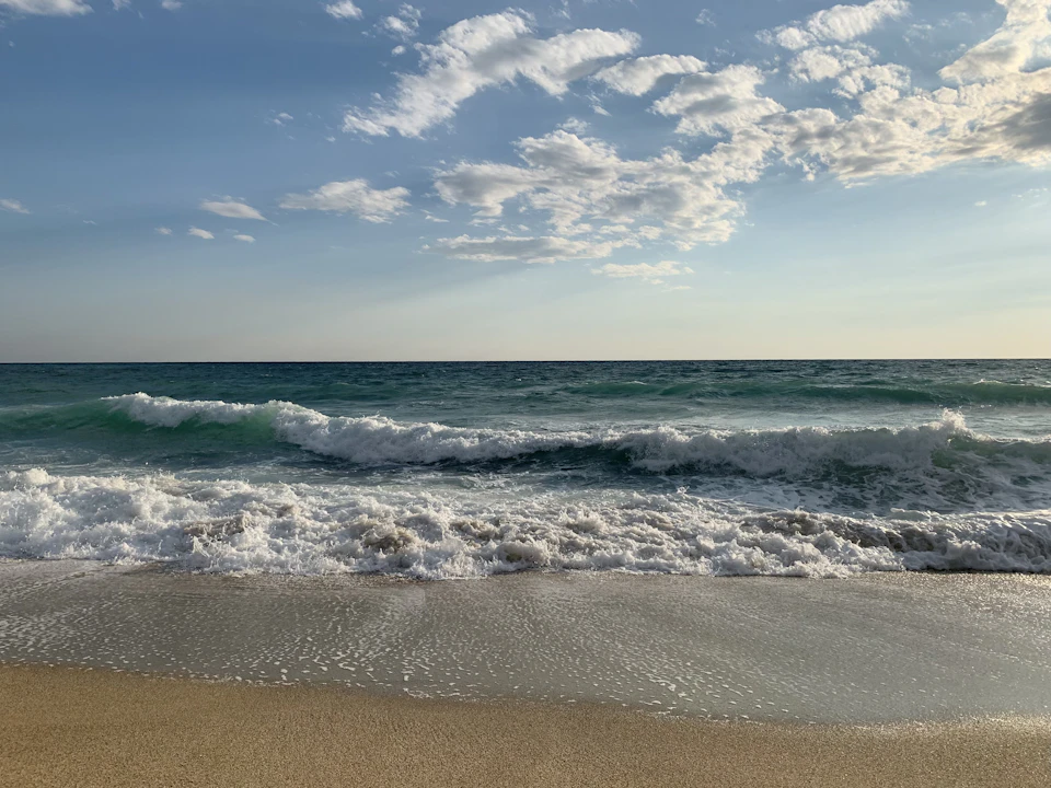 The image depicts a serene beach scene with a light brown sand surface and a deep blue ocean stretching out to meet the sky at the horizon line, where the water is breaking gently against the shoreline. The sky above is a clear blue canvas dotted with fluffy white clouds that cast soft shadows on the landscape below. The perspective of the image is from the beach, looking out towards the vast expanse of the ocean and sky.
