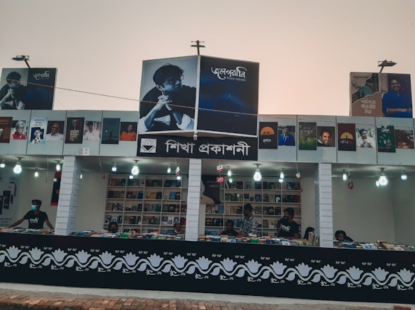 A book stall with a variety of books displayed on shelves and a counter, decorated with images of authors. Several people are present, some wearing masks, possibly indicating a public event or a book fair.