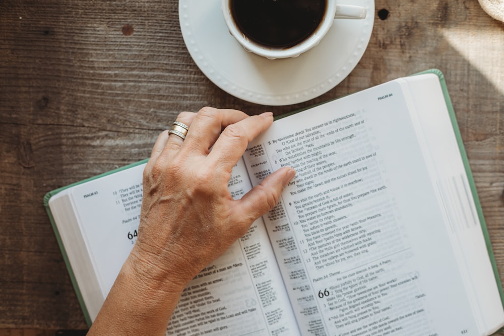 a person's hand resting on an open book next to a cup of coffee