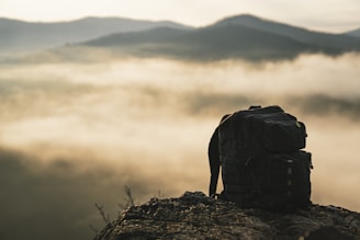 A rugged backpack sits on the edge of a rocky outcrop with misty clouds in the background. The scene conveys a sense of adventure and exploration, with distant mountains partially obscured by fog, suggesting an early morning or late afternoon time.