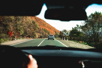 A scenic drive along a road surrounded by autumn foliage. The road is bordered by trees displaying vibrant fall colors, primarily shades of orange and green. A car is visible ahead, and the image is taken from inside another vehicle, highlighting the driver's perspective with hands on the steering wheel.