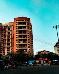A tall, multi-story orange building stands prominently against a bright blue sky. Numerous power lines crisscross the sky in front of the building. Below, trees partially obscure the ground level, where several vehicles, including auto rickshaws, are parked along the roadside. Advertising hoardings and small structures add to the urban feel of the scene.