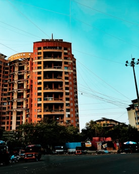 A tall, multi-story orange building stands prominently against a bright blue sky. Numerous power lines crisscross the sky in front of the building. Below, trees partially obscure the ground level, where several vehicles, including auto rickshaws, are parked along the roadside. Advertising hoardings and small structures add to the urban feel of the scene.