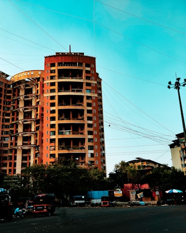 A tall, multi-story orange building stands prominently against a bright blue sky. Numerous power lines crisscross the sky in front of the building. Below, trees partially obscure the ground level, where several vehicles, including auto rickshaws, are parked along the roadside. Advertising hoardings and small structures add to the urban feel of the scene.