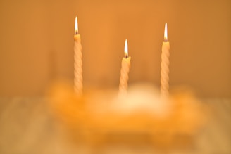 Three lit, twisted candles are arranged in a row with a soft, warm glow. The flames are steady, providing a sense of calm and tranquility. The background and foreground are blurred, accentuating the focus on the candles.