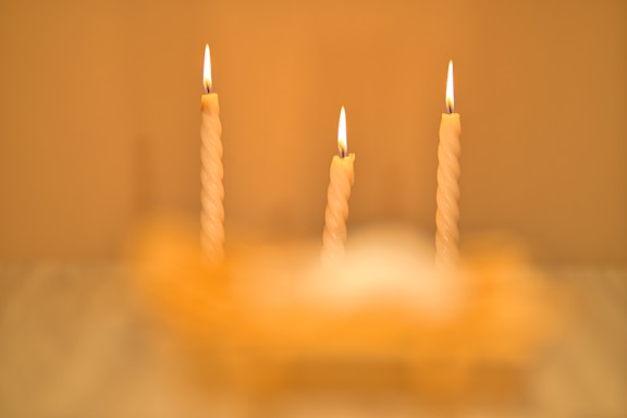 Three lit, twisted candles are arranged in a row with a soft, warm glow. The flames are steady, providing a sense of calm and tranquility. The background and foreground are blurred, accentuating the focus on the candles.