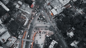An aerial view of a busy urban intersection surrounded by buildings and vegetation. Vehicles are densely packed, indicating heavy traffic. The roads form a triangular convergence, and surrounding structures vary from high-rises to lower residential and commercial buildings. The scene appears lively with visible human activity.