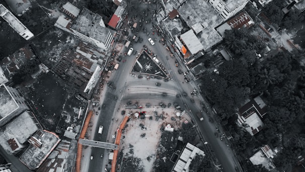An aerial view of a busy urban intersection surrounded by buildings and vegetation. Vehicles are densely packed, indicating heavy traffic. The roads form a triangular convergence, and surrounding structures vary from high-rises to lower residential and commercial buildings. The scene appears lively with visible human activity.