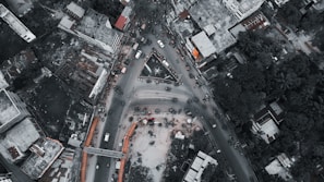 An aerial view of a busy urban intersection surrounded by buildings and vegetation. Vehicles are densely packed, indicating heavy traffic. The roads form a triangular convergence, and surrounding structures vary from high-rises to lower residential and commercial buildings. The scene appears lively with visible human activity.