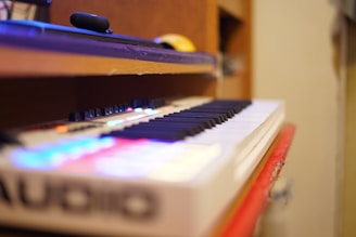 A musical keyboard with black and white keys is placed on a wooden surface. Bright LED lights illuminate part of the keyboard, adding colorful reflections. In the background, there is a blurred view of shelves and objects.