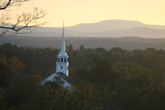A white church steeple rises above a forest of green trees, set against a backdrop of rolling hills under a golden sky. The atmosphere is calm and serene, with the sun casting a warm glow over the landscape.