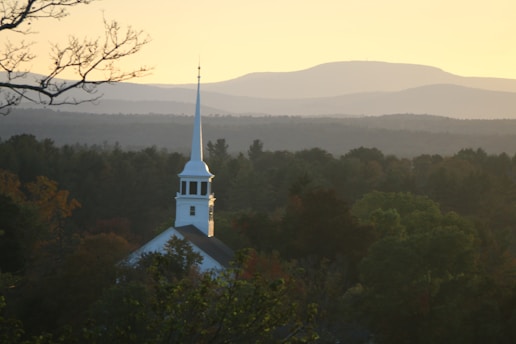 A white church steeple rises above a forest of green trees, set against a backdrop of rolling hills under a golden sky. The atmosphere is calm and serene, with the sun casting a warm glow over the landscape.