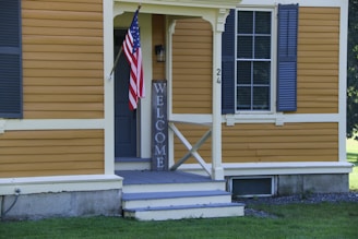 A wooden house exterior with yellow siding and white trim, featuring an American flag on a pole next to a door with the number 24. A wooden 'WELCOME' sign is leaning against the wall beside the entrance. The house has blue shutters on the window and a small set of blue and white stairs leading to the porch area. The surrounding area includes a lawn with green grass.