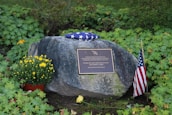 A memorial site featuring a large rock with a plaque in front. The plaque is engraved with a dedication message. On top of the rock lies a folded American flag. To the right, a small American flag is planted in the ground, and on the left, there is a pot of yellow flowers and an additional yellow flower placed on the ground. The background and surroundings are filled with green foliage and natural vegetation.