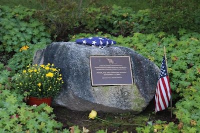 A memorial site featuring a large rock with a plaque in front. The plaque is engraved with a dedication message. On top of the rock lies a folded American flag. To the right, a small American flag is planted in the ground, and on the left, there is a pot of yellow flowers and an additional yellow flower placed on the ground. The background and surroundings are filled with green foliage and natural vegetation.