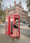 A woman wearing a black top, pink skirt, sunglasses, and a hat is posing by two iconic red British telephone booths. The background features a large, historic building with intricate architecture and multiple chimneys, and there are some trees nearby.