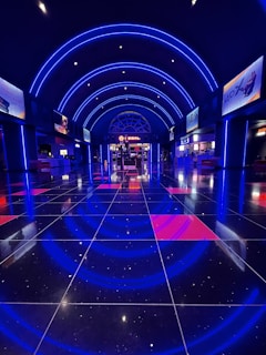A spacious movie theater lobby features sleek, shiny black floor tiles reflecting vibrant blue neon lights. The ceiling is adorned with symmetrical arched blue lighting extending down the walls. Multiple illuminated screens and advertisements line the walls, while the entrance to a theater can be seen in the background with a bright circular light overhead.