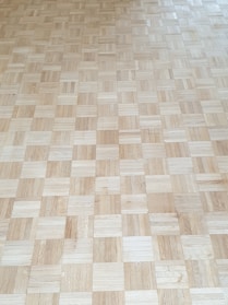 A wooden parquet floor is composed of square tiles arranged in a checkered pattern. The tiles vary slightly in color and grain, creating a natural and textured appearance.