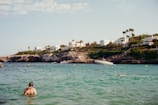 A scenic coastal area features turquoise waters with a swimmer in the foreground wearing snorkeling gear. In the background, white modern houses are perched on rocky cliffs surrounded by greenery. A boat is anchored near the shore, and several people are swimming in the water. The sky is mostly clear with a few clouds.