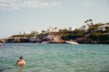 A scenic coastal area features turquoise waters with a swimmer in the foreground wearing snorkeling gear. In the background, white modern houses are perched on rocky cliffs surrounded by greenery. A boat is anchored near the shore, and several people are swimming in the water. The sky is mostly clear with a few clouds.