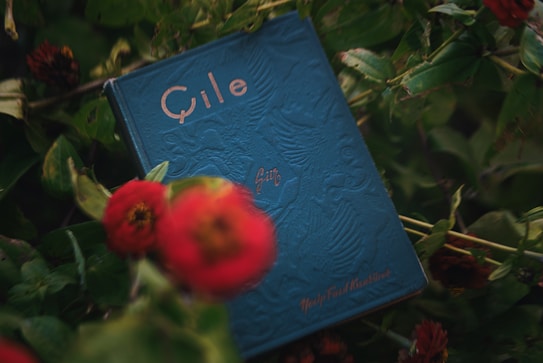 A blue book with embossed designs, featuring the title written in a stylized font, is surrounded by lush green foliage and red flowers. The book cover has an intricate pattern with faded text visible, placed against a natural backdrop creating a harmonious blend of nature and literature.