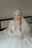 A bride with blonde hair sits gracefully, wearing an elegant white lace wedding dress adorned with intricate patterns. She accessorizes with a delicate, sparkling tiara that complements the veil draping over her shoulders. The expression on her face is serene and composed. The backdrop features a soft, wavy patterned wallpaper and a cushioned headboard.