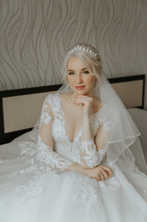 A bride with blonde hair sits gracefully, wearing an elegant white lace wedding dress adorned with intricate patterns. She accessorizes with a delicate, sparkling tiara that complements the veil draping over her shoulders. The expression on her face is serene and composed. The backdrop features a soft, wavy patterned wallpaper and a cushioned headboard.