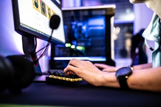 A person is using a desktop computer, focusing on the keyboard and monitor. The setup includes a microphone and several peripheral devices visible, suggesting a workstation or gaming environment. The hands are actively typing, and there is ambient lighting reflecting off the computer case.