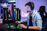 A young person is intensely focused on a computer screen while wearing a headset with a microphone. They are using a gaming keyboard and mouse, sitting in a gaming chair. The room has cool lighting, with a visible PC tower.