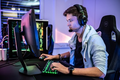 A young person is intensely focused on a computer screen while wearing a headset with a microphone. They are using a gaming keyboard and mouse, sitting in a gaming chair. The room has cool lighting, with a visible PC tower.